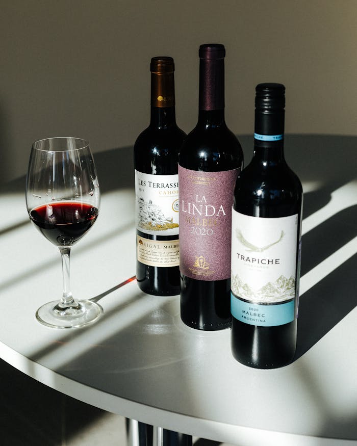 Three red wine bottles and a filled wine glass on a table in natural light.