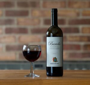 Barolo Riserva 2015 bottle with wine glass on a wooden table against a brick wall.